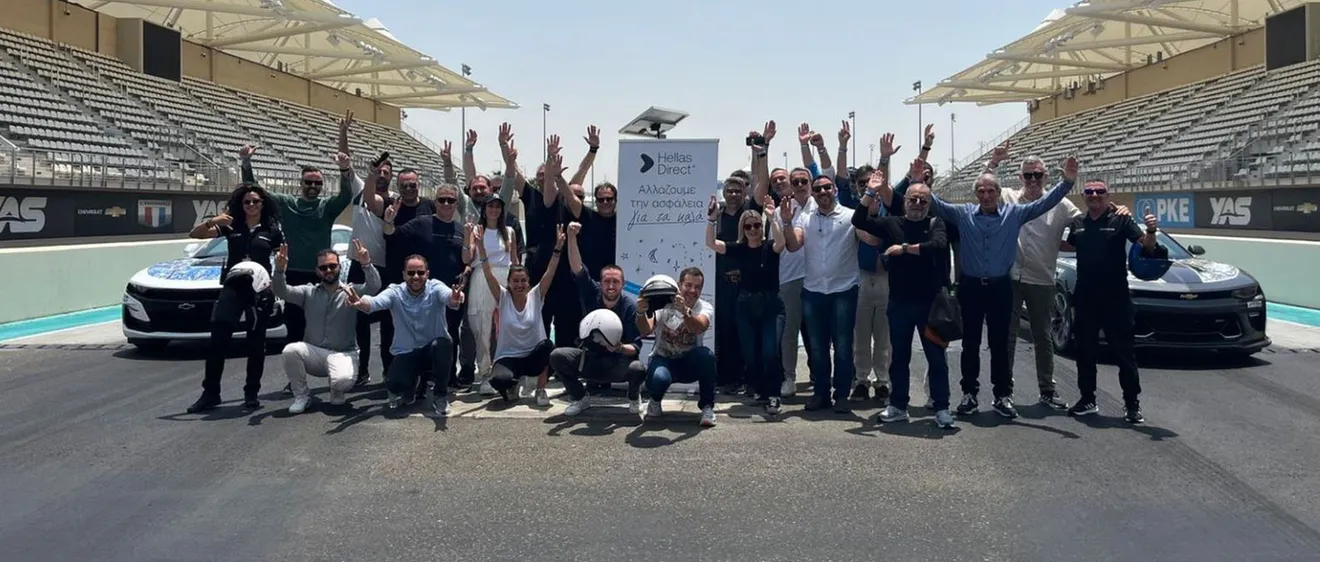 Ομάδα ανθρώπων σε πίστα αγώνων, πανηγυρίζουν μπροστά από πανό "Hellas Direct", ανάμεσα σε δύο αυτοκίνητα.
