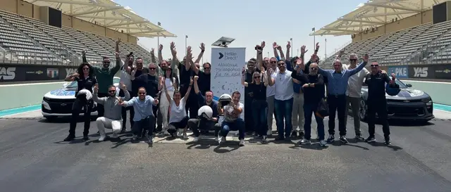 Ομάδα ανθρώπων σε πίστα αγώνων, πανηγυρίζουν μπροστά από πανό "Hellas Direct", ανάμεσα σε δύο αυτοκίνητα.