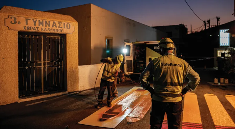 Δύο εργάτες με κράνη βάφουν διαβάσεις σε δρόμο, μπροστά από το 3ο Γυμνάσιο Χώρας Καλύμνου, κατά τη διάρκεια της νύχτας.