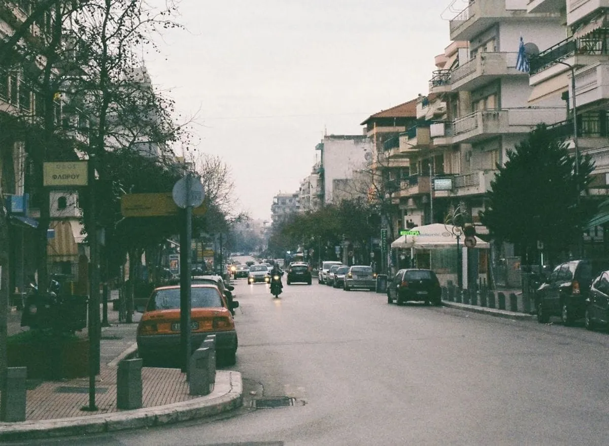 εικόνα δρόμου πόλης, άσφαλτος στη μέση, αυτοκίνητα παρκαρισμένα και εν κινήσει, περίπτερο και κτήρια δεξιά αριστερά